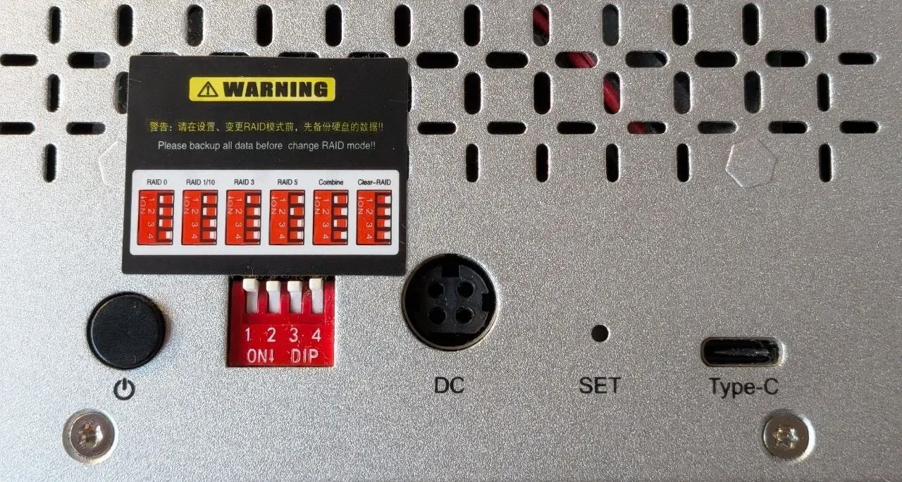 Parte trasera de la carcasa Yottamaster de 4 bahías antes del cambio: los cuatro interruptores DIP están hacia arriba, en OFF. Encima, la etiqueta de advertencia con la combinación de cada modo RAID; abajo, el conector DC, el orificio del botón SET y el puerto USB-C.